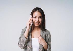 ung Lycklig asiatisk företag kvinna med lång hår i kostym använder sig av mobil telefon på grå bakgrund foto