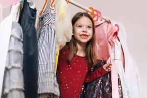 leende liten flicka med lång mörk hår i röd klänning bland henne skön klänningar i garderob i barns rum på Hem foto