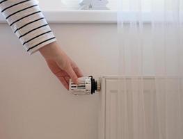 kvinna reglerar temperatur på Hem med de uppvärmning termostat till spara energi, närbild med hand. temperatur kontrollera ventil på radiator. vit värmare på vägg med ridå i modern lägenhet. foto
