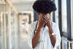 ledsen och deprimerad ung afrikansk amerikan kvinna läkare i vit enhetlig stående i korridor foto