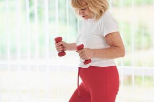 positiv senior kvinna i sportigt kläder inomhus på dagtid håller på med övningar med hantlar foto