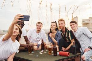 flicka håller på med Foto när människor spelar kort spel. grupp av ung människor i tillfällig kläder ha en fest på taket tillsammans på dagtid