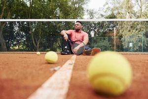 sitter nära netto och tar en ha sönder. afrikansk amerikan man i rosa skjorta sitter med tennis racket på de domstol utomhus foto