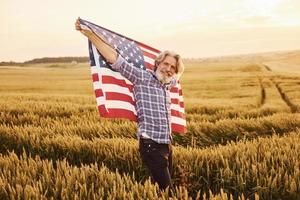 innehav USA flagga i händer. patriotisk senior eleganta man med grå hår och skägg på de jordbruks fält foto