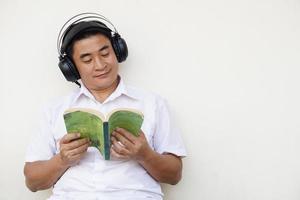 asiatisk man läser bok och bär hörlurar lyssna till mjuk musik till göra koncentration i läsning. begrepp, fri tid, tidsfördriv, hobby, koppla av med musik. foto