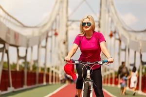 skön flicka med blond hår lyckligt ridning cykel. porträtt av ung lady i j solglasögon har roligt, ridning på cykel längs stad gator foto