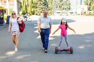 liten leende flicka inlärning till rida en hoverboard med henne föräldrar utomhus foto