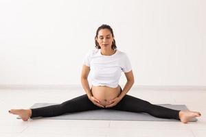 ung flexibel gravid kvinna håller på med gymnastik på matta på de golv på vit bakgrund. de begrepp av framställning de kropp för lätt förlossning foto