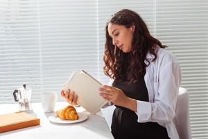 sida se av Lycklig ung skön kvinna läsning bok medan har morgon- frukost med kaffe och croissanter på bakgrund av persienner. Bra morgon- begrepp och trevlig lunch ha sönder foto