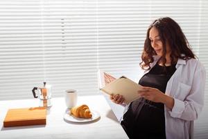 sida se av Lycklig ung skön kvinna läsning bok medan har morgon- frukost med kaffe och croissanter på bakgrund av persienner. Bra morgon- begrepp och trevlig lunch ha sönder foto