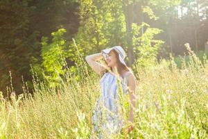 Lycklig ung kvinna med lång hår i hatt och klänning gående genom de sommar skog på en solig dag. sommar glädje begrepp foto