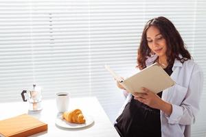 sida se av Lycklig gravid kvinna läsning bok medan har morgon- frukost med kaffe och croissanter på bakgrund av persienner. Bra morgon- begrepp och trevlig lunch ha sönder foto