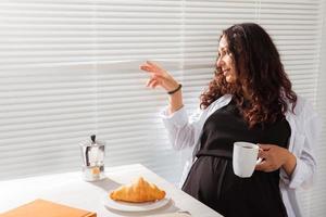 glad gravid ung skön kvinna utseende genom persienner under henne morgon- frukost med kaffe och croissanter. begrepp av Bra morgon- och väntar för möte med en bebis. copy foto