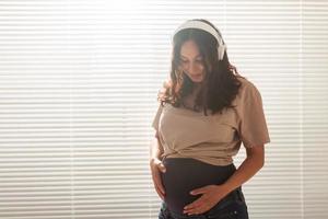 Lycklig gravid kvinna med lockigt hår lyssnande musik i hörlurar, kopia Plats. foto