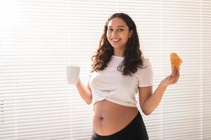 skön gravid kvinna innehav croissant och kopp av kaffe i henne händer under morgon- frukost. begrepp av Bra hälsa och positiv attityd medan förväntar sig bebis foto