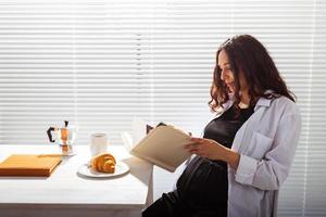 sida se av Lycklig gravid kvinna läsning bok medan har morgon- frukost med kaffe och croissanter på bakgrund av persienner. Bra morgon- begrepp och trevlig lunch ha sönder foto