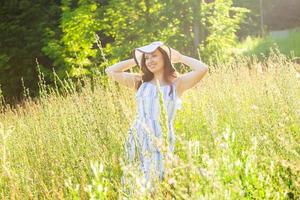 Lycklig ung kvinna med lång hår i hatt och klänning gående genom de sommar skog på en solig dag. sommar glädje begrepp foto