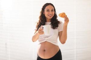 skön gravid kvinna innehav croissant och kopp av kaffe i henne händer under morgon- frukost. begrepp av Bra hälsa och positiv attityd medan förväntar sig bebis foto