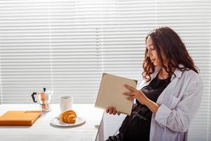 sida se av Lycklig gravid kvinna läsning bok medan har morgon- frukost med kaffe och croissanter på bakgrund av persienner. Bra morgon- begrepp och trevlig lunch ha sönder foto