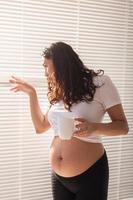 nyfiken ung skön gravid kvinna dricka te och ser genom de persienner på de fönster. begrepp av glädje och Bra Nyheter medan väntar för bebis foto
