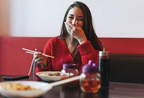 skön leende ung asiatisk kvinna i röd kläder äter asiatisk mat med bambu ätpinnar i de kinesisk vietnamese restaurang foto