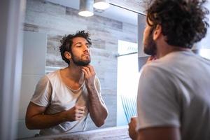 reflexion av stilig man med skägg ser på spegel och rörande ansikte i badrum grooming foto