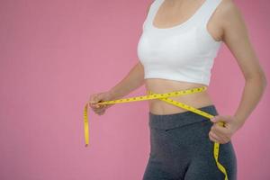 smal kvinna i sportkläder åtgärder henne midja använder sig av tejp mäta på rosa bakgrund. diet kvinna och tappa bort vikt planen foto