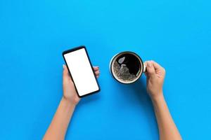 kvinna händer innehav svart mobil telefon med tom vit skärm och råna av kaffe. attrapp bild med kopia Plats. topp se på blå bakgrund, platt lägga foto