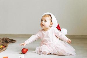 söt liten bebis flicka i rosa klänning med tutu kjol och santa hatt med närvarande gåva låda Sammanträde på golv i rum med jul leksaker på Hem, Lycklig ny år foto