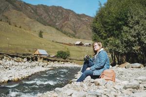 skön leende blond ung kvinna resande med Foto kamera i händer nära de berg flod