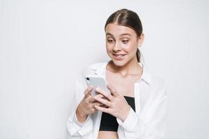 emotionell porträtt av Lycklig tonåring flicka med lång hår i vit skjorta använder sig av mobil telefon på vit bakgrund foto