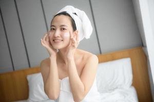 Lycklig ung asiatisk kvinna applicering ansikte lotioner medan bär en handduk och rörande henne ansikte. dagligen smink och hudvård foto