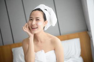 Lycklig ung asiatisk kvinna applicering ansikte lotioner medan bär en handduk och rörande henne ansikte. dagligen smink och hudvård foto