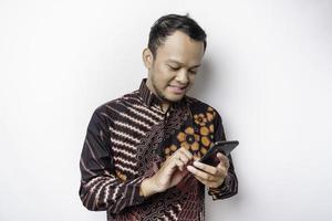 en porträtt av en Lycklig asiatisk man bär batik skjorta och innehav hans telefon, isolerat förbi vit bakgrund foto