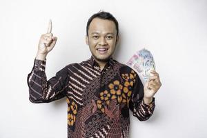 en Lycklig ung asiatisk man är bär batik skjorta, pekande på de kopia Plats på topp av honom medan innehav pengar i indonesiska rupiah isolerat förbi vit bakgrund foto