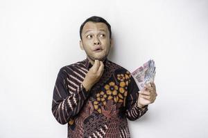 en omtänksam ung asiatisk man är bär batik skjorta och innehav kontanter pengar i indonesiska rupiah isolerat förbi vit bakgrund foto