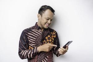 en porträtt av en Lycklig asiatisk man bär batik skjorta och innehav hans telefon, isolerat förbi vit bakgrund foto