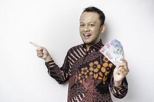 en Lycklig ung asiatisk man är bär batik skjorta, pekande på de kopia Plats bredvid honom medan innehav pengar i indonesiska rupiah isolerat förbi vit bakgrund foto