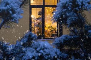 mysigt festlig fönster av de hus utanför med de värma ljus av fe- lampor kransar inuti - fira jul och ny år i en värma Hem. jul träd, bokeh, snö på tall träd och snöfall foto
