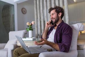 ung attraktiv leende kille är bläddring på hans bärbar dator och talande åh mobil telefon, Sammanträde på Hem på de mysigt soffa, bär tillfällig utrusta. frilans företag arbete från Hem begrepp foto