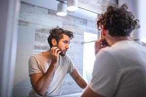 morgon- hygien, stilig man i de badrum ser i spegel. reflexion av stilig man med skägg ser på spegel och rörande ansikte i badrum grooming foto