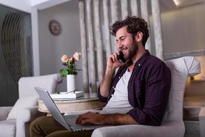 ung stilig man framställning video ring upp med vänner medan Sammanträde på soffa på hans modern hem.koncept av Lycklig företag människor. företag man på Hem kontroll statistik på mobil telefon och bärbar dator foto