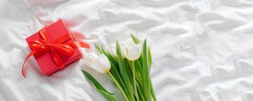 vit tulpaner, röd gåva låda på säng. fortfarande liv, minimal begrepp. kvinnors dag, mödrar dag Semester. baner för webb sida foto