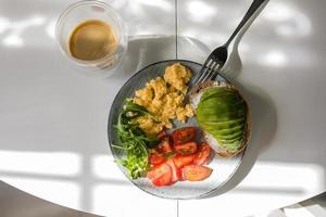 tallrik med krypterade ägg, ruccola, tomat, hela vete rostat bröd med avokado, kopp av kaffe på tabell. foto