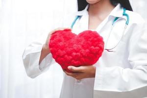 en skön asiatisk kvinna läkare innehav en stor röd hjärta. i de behandling rum. sjukhus patient vård begrepp foto