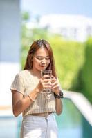 asiatisk kvinna innehar en glas av vatten från dricka på utomhus- fält med bokeh grön baner bakgrund. foto