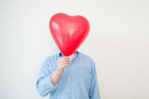 oigenkännlig man innehav hjärta ballong i främre av hans ansikte mot vit vägg. valentines bakgrund med copy foto