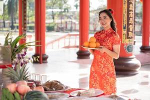 skön ung asiatisk kvinna leende lyckligt bärande traditionell Cheongsam qipao klänning innehav färsk apelsiner be för bäst önskar välsignelse och Bra tur i kinesisk buddist tempel. foto