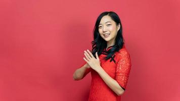 asiatisk kvinna bär traditionell Cheongsam qipao klänning med gest av congratulation isolerat på röd bakgrund. Lycklig kinesisk ny år foto