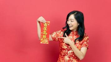 skön kvinna bär traditionell Cheongsam qipao kostym innehav och pekande välsignelse förmögenhet kort isolerat på röd bakgrund. kinesisk text betyder bra tur bra vinst foto